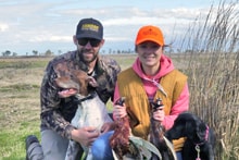 Man and woman kneeling in a field with a black Labrador and two harvested pheasants