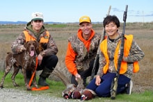 Three hunters in blaze orange kneeling with a German Wirehaired Pointer and harvested pheasants in a green field