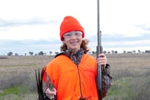 Young woman hunter in full camo and cap holding a shotgun and harvested pheasant in a grassy field