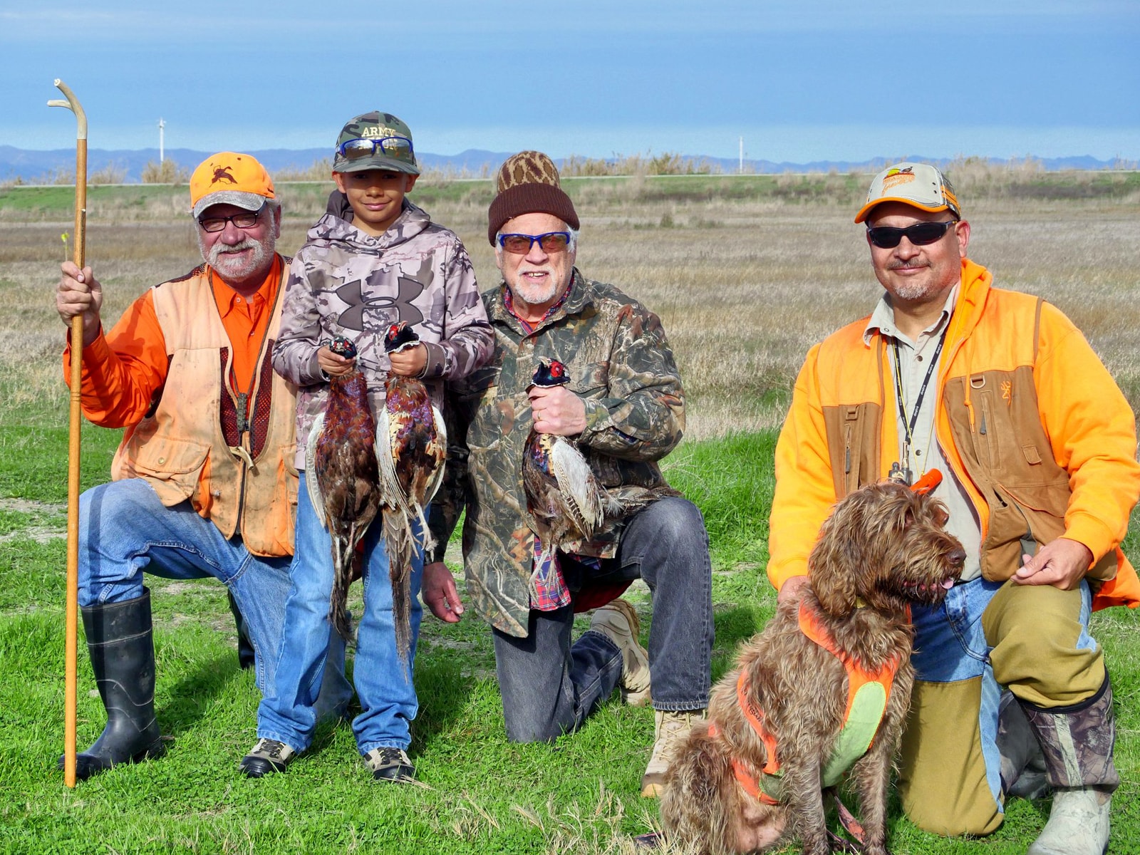 Woman hunter in camo vest and blaze orange holding two harvested rooster pheasants in a green field