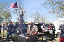 Man in a mask tending a large outdoor BBQ grill with smoke rising, surrounded by participants at picnic tables under an American flag at the 2022 Veterans Upland Bird Hunt