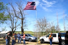 Group of hunters in orange vests gathered around a large American flag flying on a tall pole at the 2022 Veterans Upland Bird Hunt