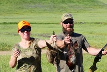 Man and woman in camo each holding harvested pheasants and a shotgun in a green field at the 2022 Veterans Upland Bird Hunt