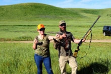 Man in camo and woman in blaze orange cap standing together in a green field — man demonstrating shotgun handling technique