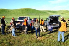 Group of hunters in blaze orange gathered around trucks in a green rolling-hills field for a pre-hunt briefing at the 2022 Veterans Upland Bird Hunt