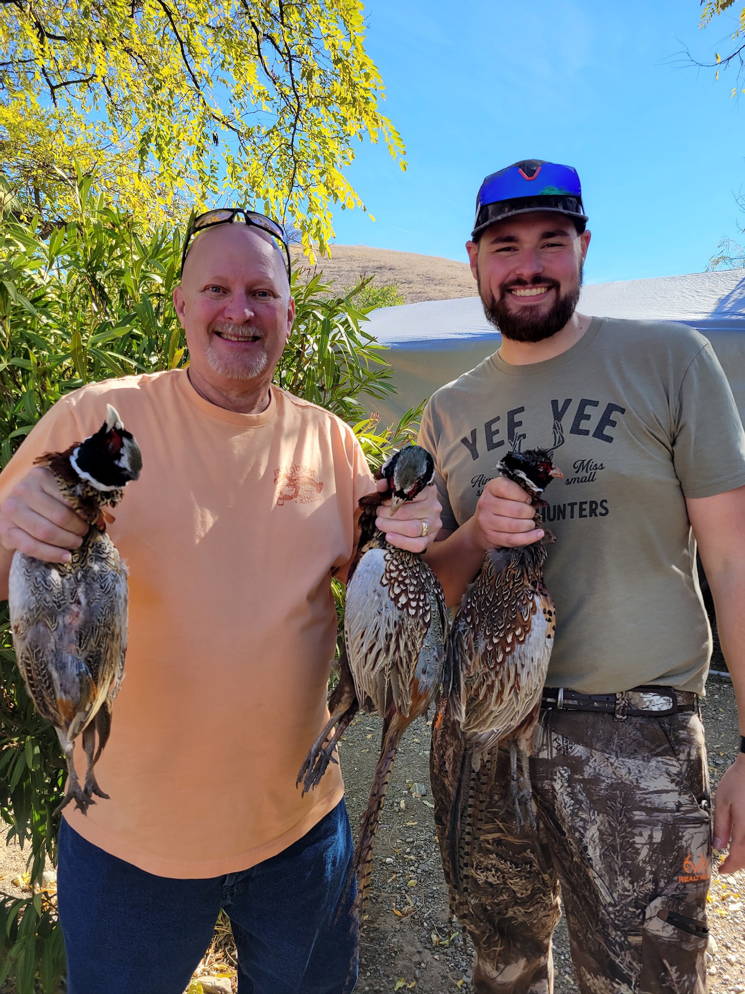 Two hunters — one in a peach shirt, one in a Yee Yee camo shirt — each holding harvested pheasants under a yellow-leafed tree at the 2023 Veterans and First Responders Hunt