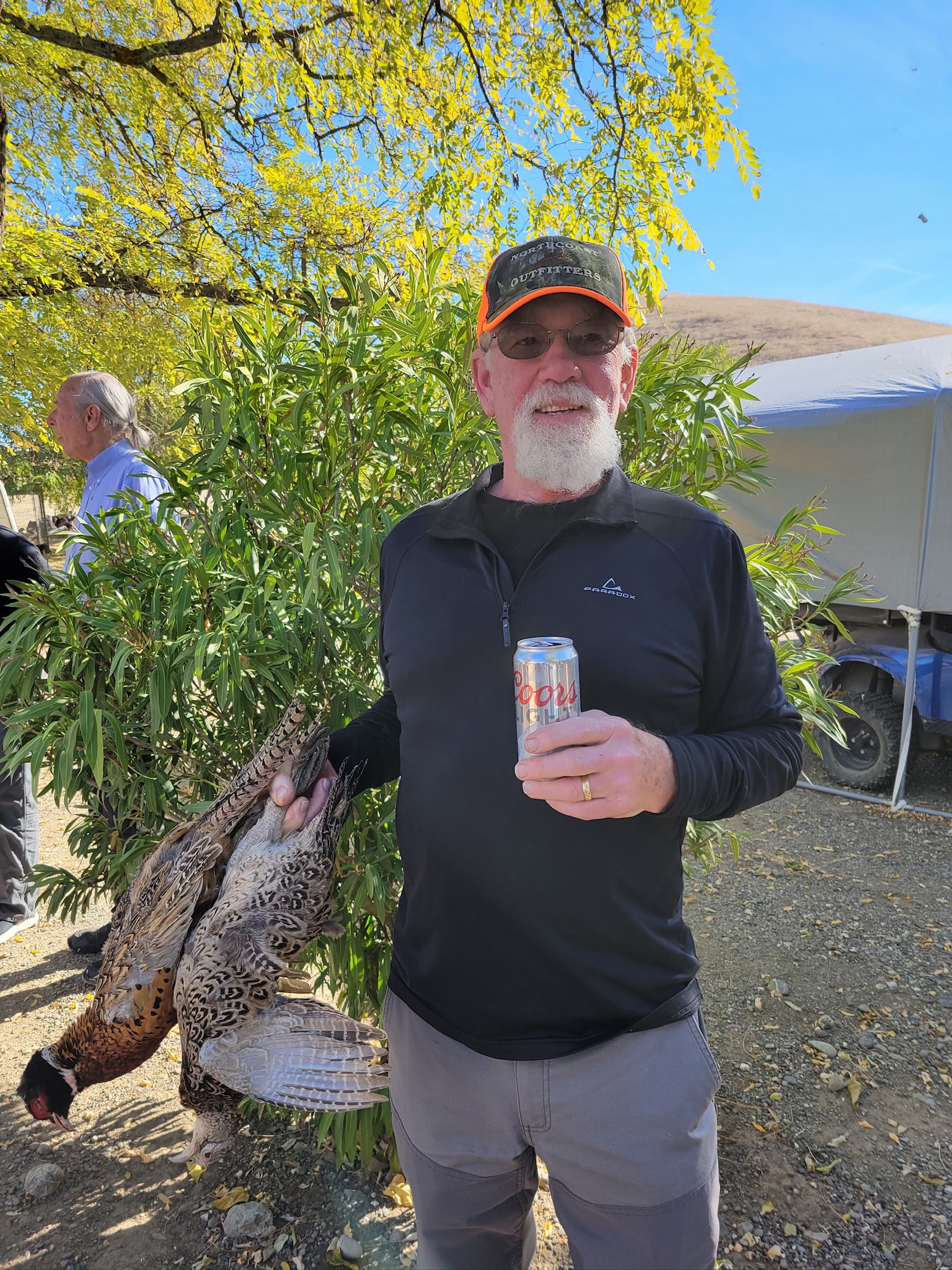 Hunter in a black pullover and North Coast Outfitters cap holding a harvested pheasant and a Coors Light under a yellow-leafed tree at the 2023 Veterans and First Responders Hunt