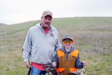 Adult mentor and young hunter in blaze orange vest standing together in a green rolling-hills field at the 2023 Youth Mentor Hunt