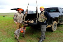 Two hunters — one in camo with a German Shorthaired Pointer, one in a blaze orange vest — standing at the tailgate of a truck with their pheasant harvest in a green field at the 2024 R3 Upland Bird Hunt