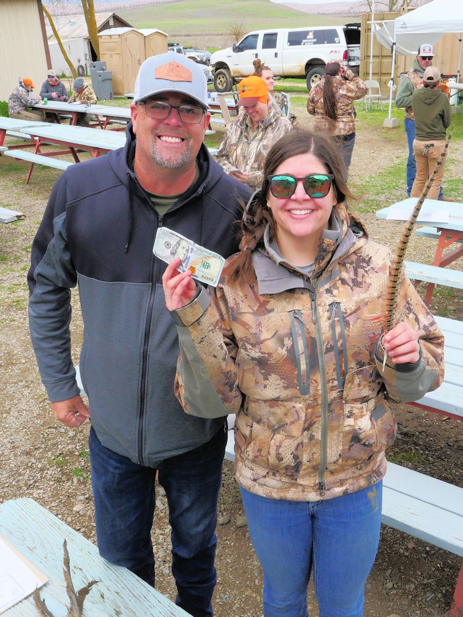 Chapter member and woman hunter in camo smiling together — she holds a $100 bill prize and a pheasant tail feather — with other participants and picnic tables in the background at the 2024 Women's Upland Bird Hunt