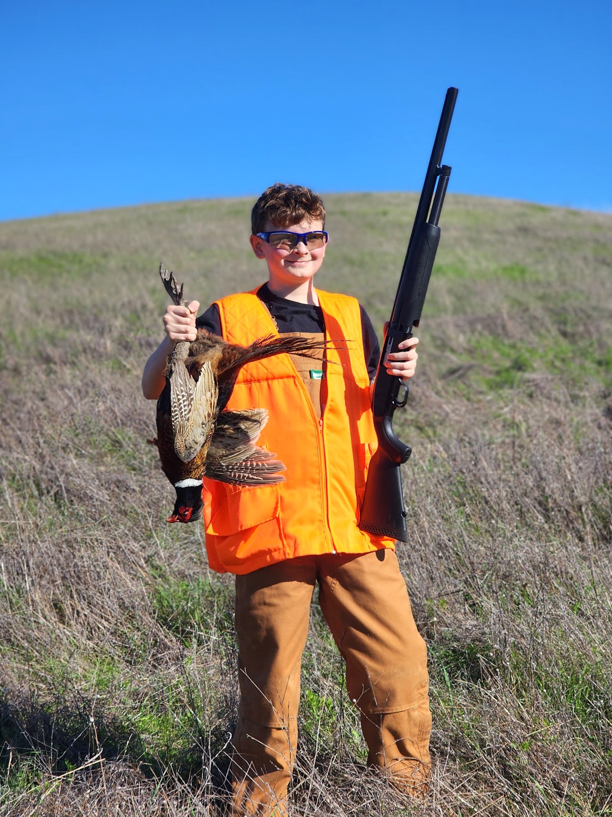 Young boy in a blaze orange vest holding a harvested pheasant by the tail and a black shotgun with a green hillside behind him at the 2024 Youth Mentor Hunt