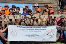 Large group of youth hunters and mentors in blaze orange vests posing in front of the Pheasants Forever Yolo County chapter banner with their pheasant harvest at the 2024 Youth Mentor Hunt