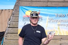 Teenage boy in a black shirt and NRA cap holding a prize in front of the Pheasants Forever Yolo County banner at the 2024 Youth Mentor Hunt