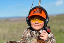 Young boy in blaze orange cap, yellow shooting glasses, and camo gear holding a harvested pheasant close-up at the 2024 Youth Mentor Hunt