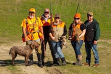 Group of five hunters in orange vests posing with a dog and harvested pheasants in a field