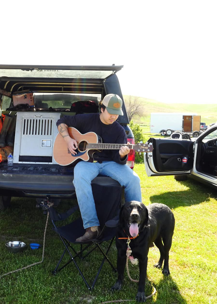 Hunter playing acoustic guitar at the tailgate of a truck with a black Labrador retriever beside him