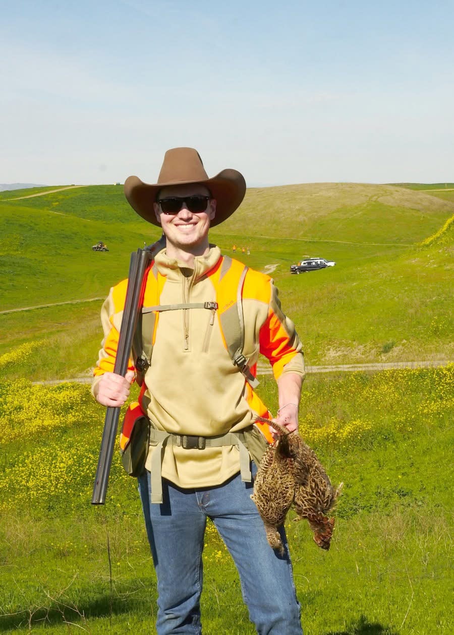Hunter in orange vest and cowboy hat holding a shotgun and harvested pheasants on a green hillside