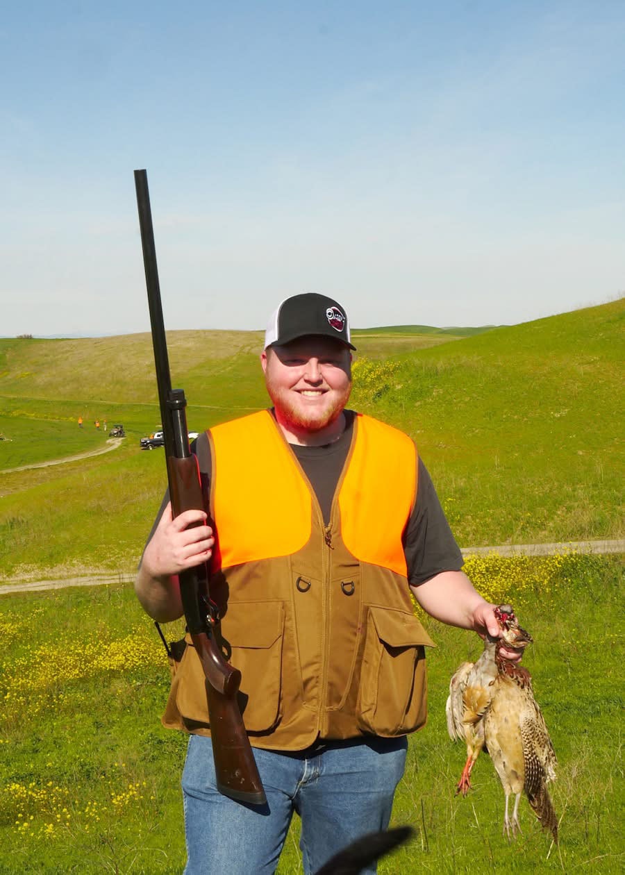 Smiling hunter in orange vest holding a shotgun and a harvested pheasant on a green hillside