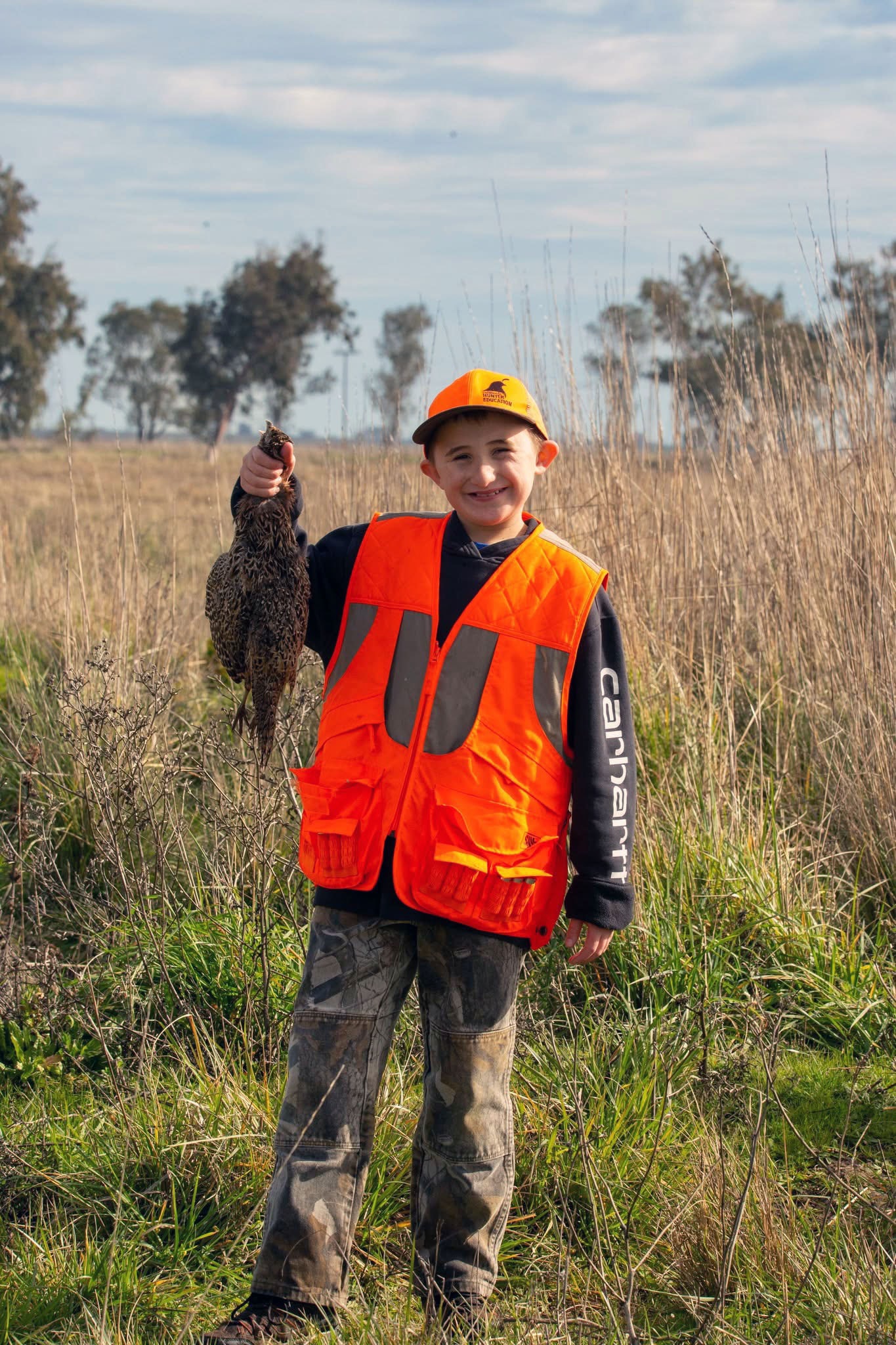 Smiling boy in orange vest holding a pheasant in a grassy field
