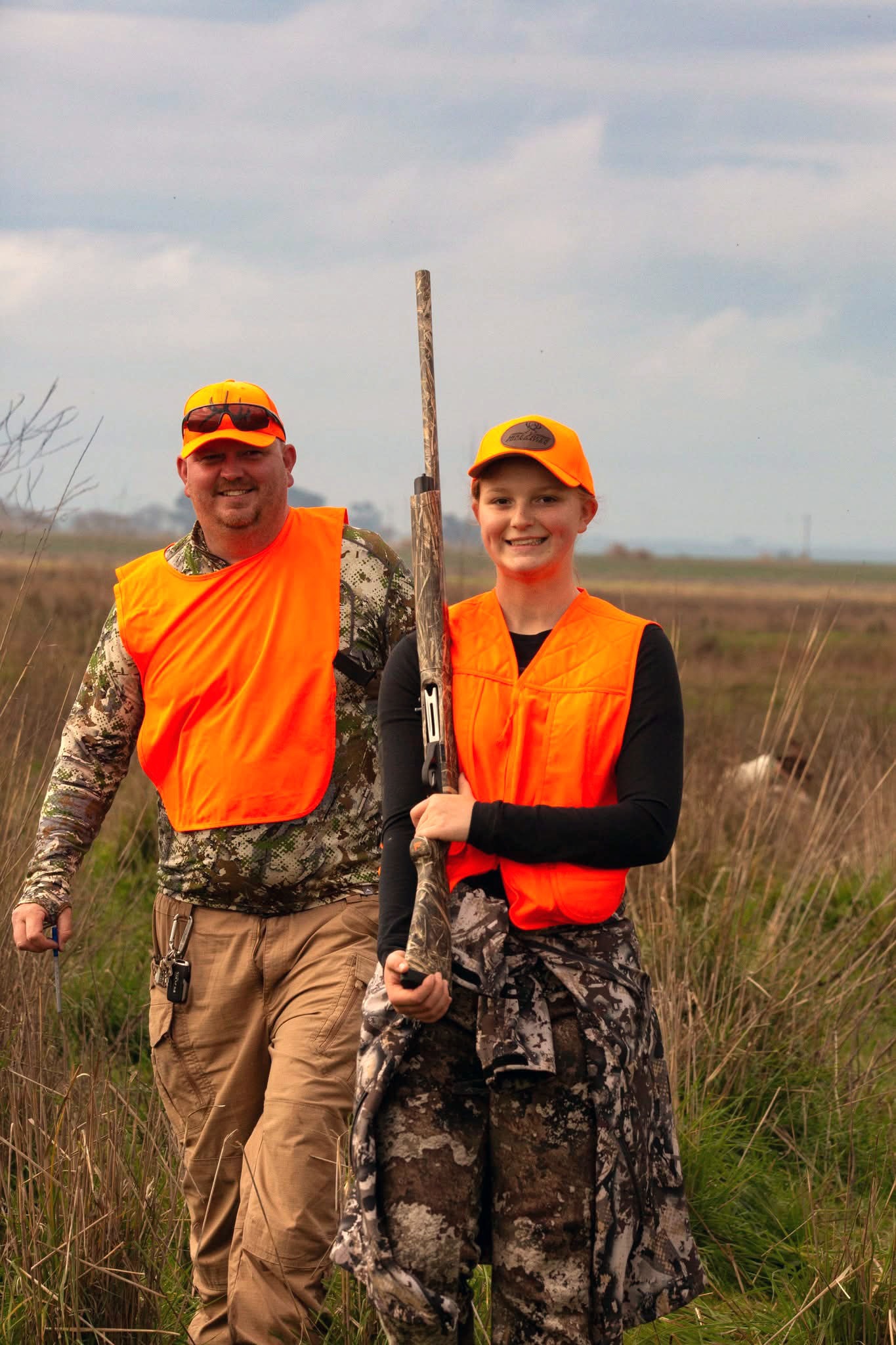 Young girl in orange vest proudly holding a pheasant with her mentor beside her