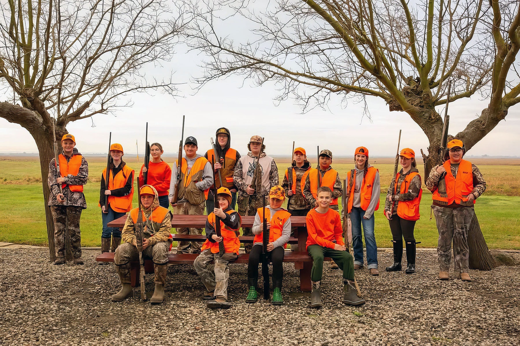 Large group of youth hunters in orange vests posing under bare trees at a picnic table