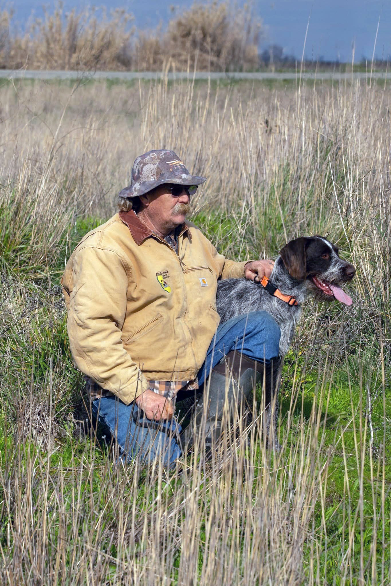 Hunter kneeling in tall grass with a German wirehaired pointer