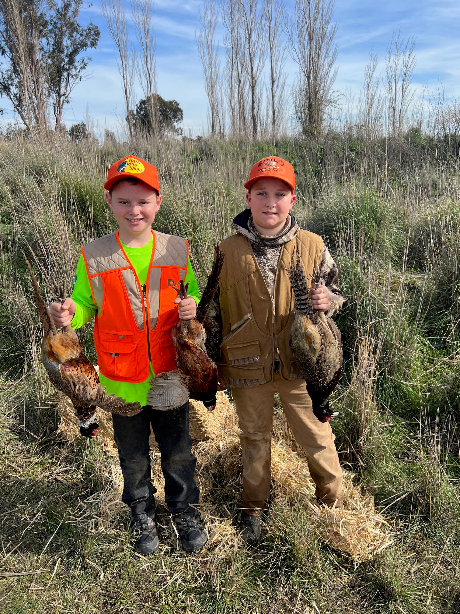Two young boys in orange vests holding harvested pheasants in a field