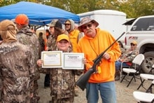 Hunter in orange vest holding a certificate and shotgun with a young hunter beside him at the awards area