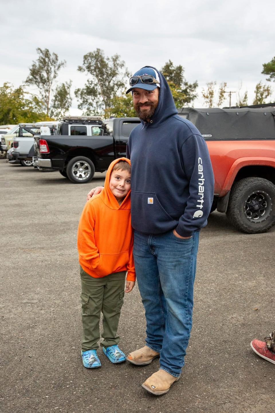 Father in a Carhartt hoodie posing with his young son in an orange hoodie in the parking area before the hunt