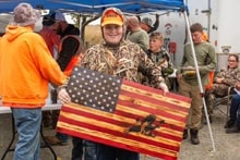 Hunter in camouflage and orange hat proudly holding a handcrafted wooden American flag at the Youth Upland Bird Hunt event