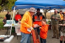 Mentor in orange vest presenting a wooden flag to a young hunter under a blue canopy with other hunters looking on