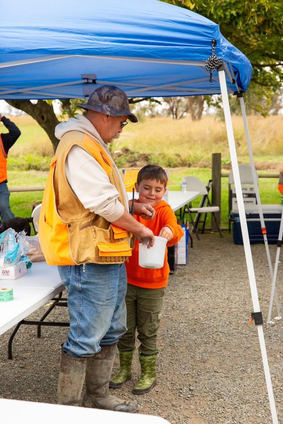 Mentor in orange vest helping a young boy in an orange hoodie under a blue canopy tent at the hunt event