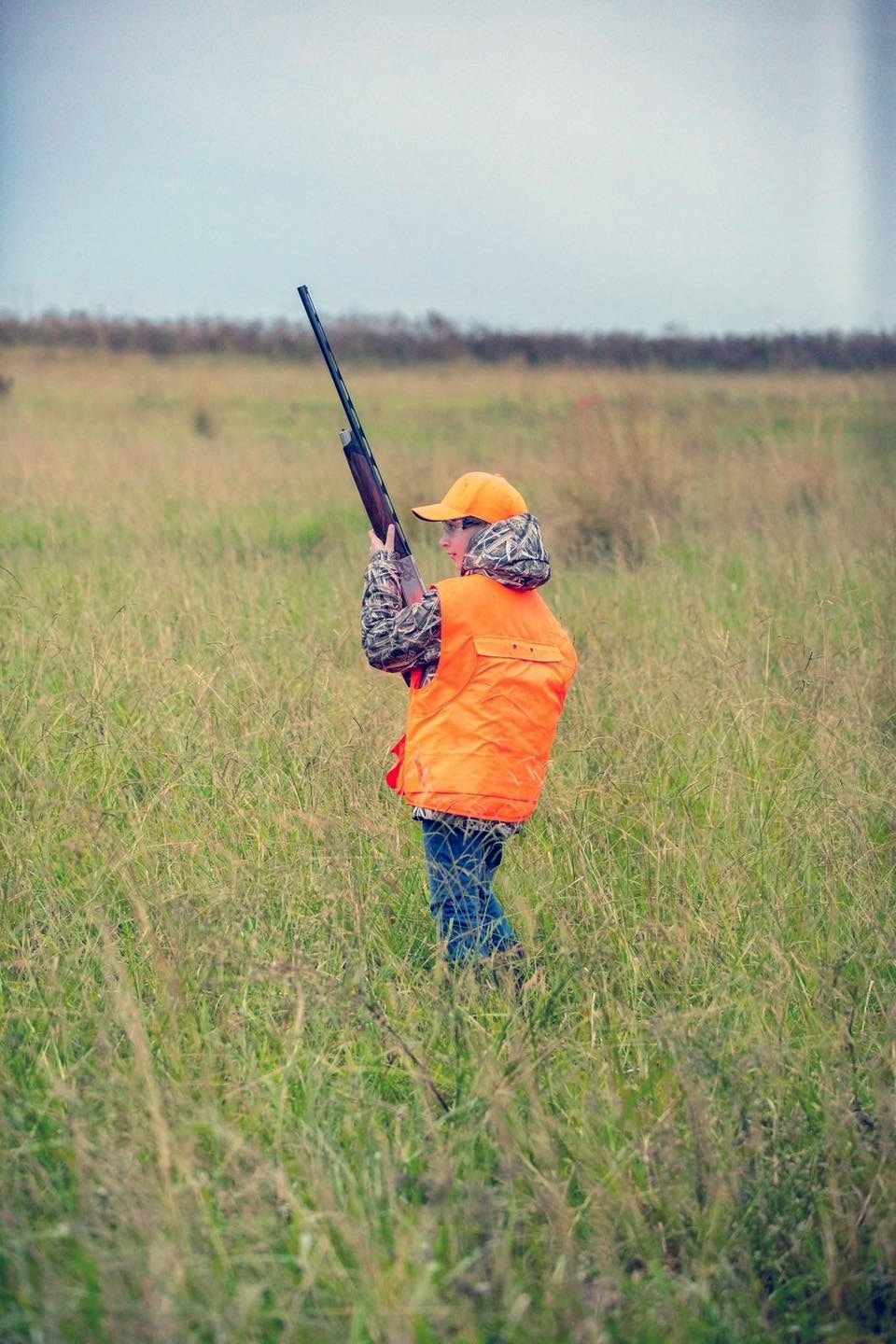Young hunter in orange vest and camo jacket standing in a green field with a shotgun raised, ready to shoot